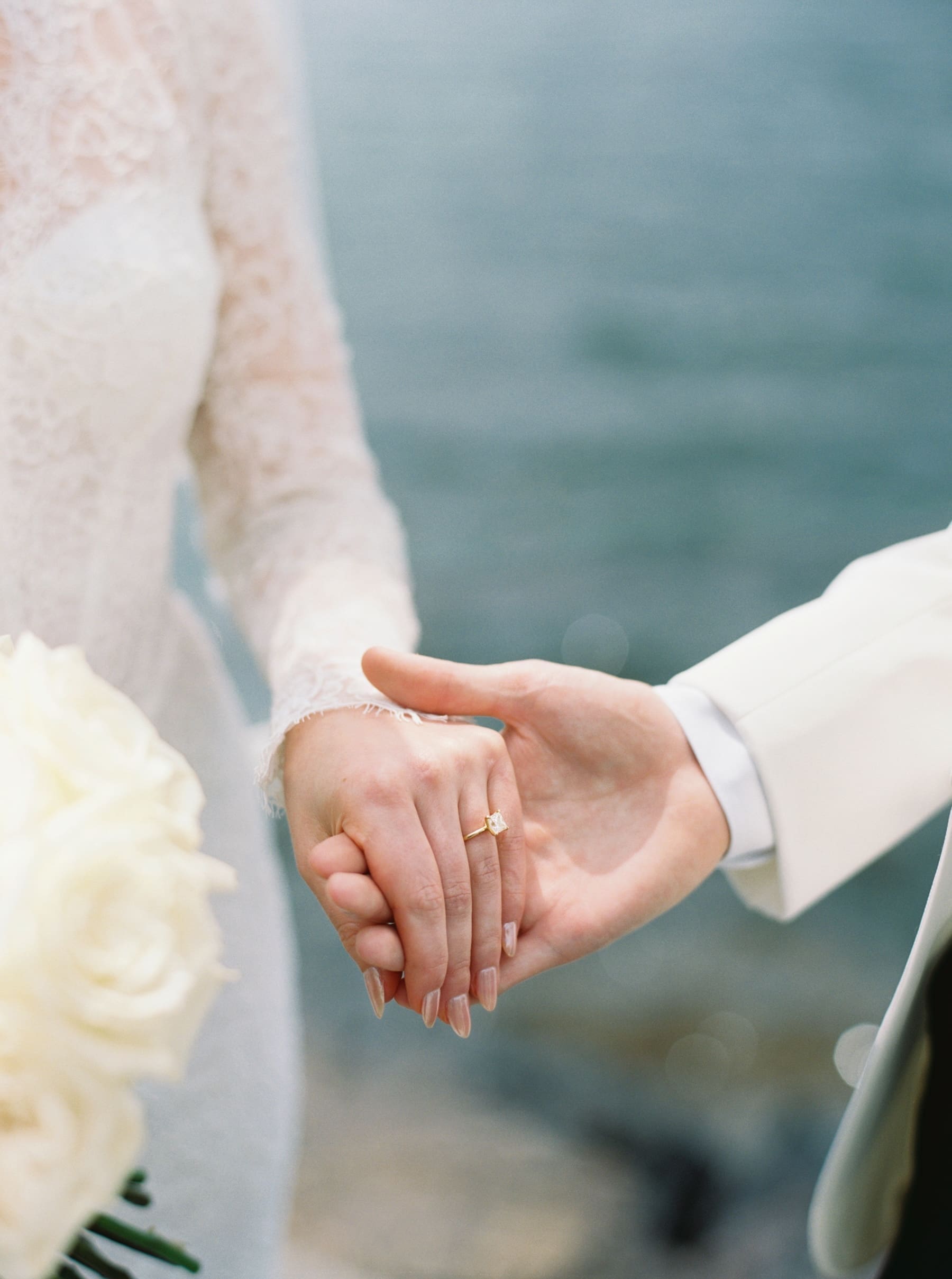 close up photo of groom holding brides hand showing off diamond ring with lake in the background 